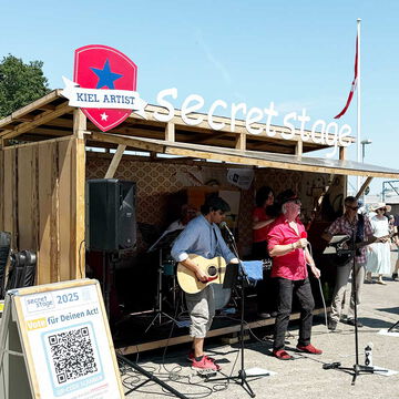 Musiker mit Gitarren vor der secret stage mit Kiel Artist Schild und Voting-Aufsteller, Kieler Woche 2025