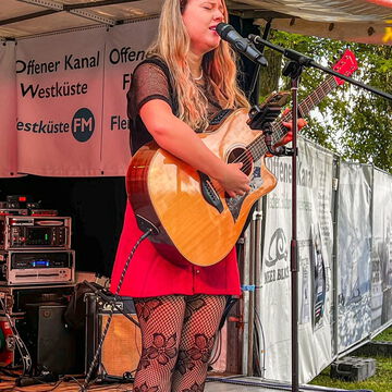 Sängerin mit Akustikgitarre im roten Rock auf der Bühne des Offenen Kanals, secret stage, Kieler Woche 2024